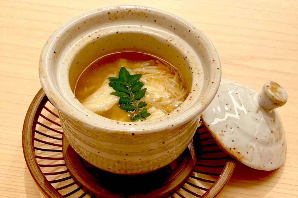 An exquisite chawanmushi or clear Japanese soup served in an artisanal ceramic bowl with a lid, garnished with fresh greens, highlighting the refined omakase dining experience at Sushi Sakuta.