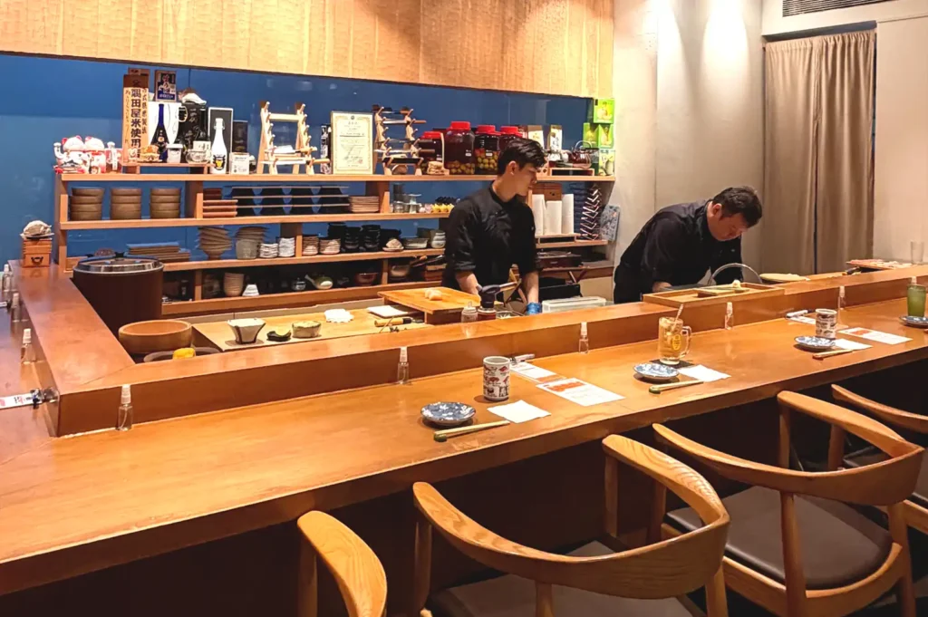 Inside view of a minimalist Japanese dining bar featuring a polished wooden counter, minimalist wooden armchairs, and chefs preparing meals behind shelves.