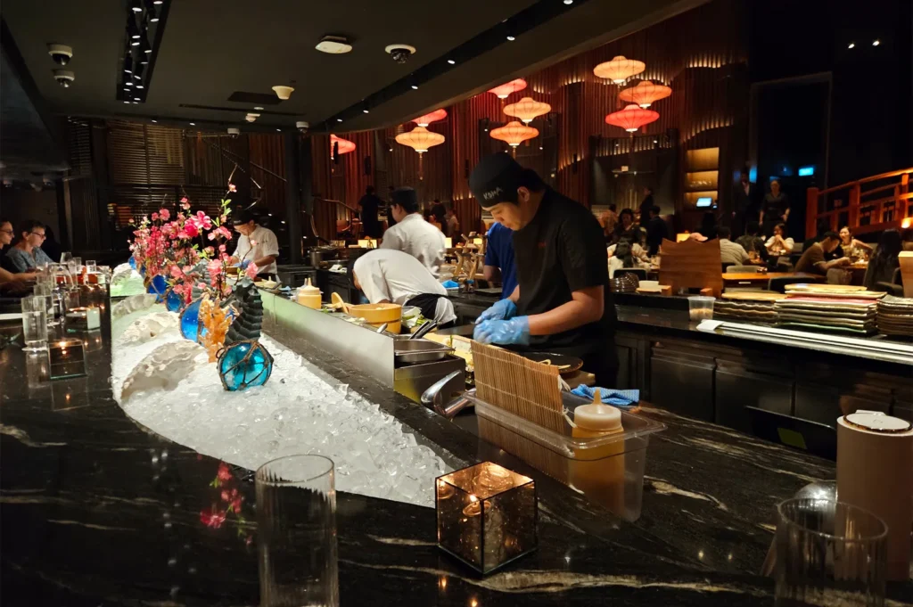 The elegant, dimly lit sushi bar counter at KOMA Singapore, Marina Bay Sands, with chefs preparing fresh ingredients on ice.