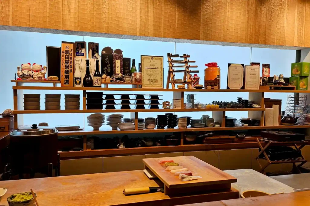 A detailed view of a minimalist Japanese sushi bar counter featuring wooden shelves stocked with sake bottles, ceramic tableware, and certificates, with fresh slices of fish ready on a wooden cutting board.