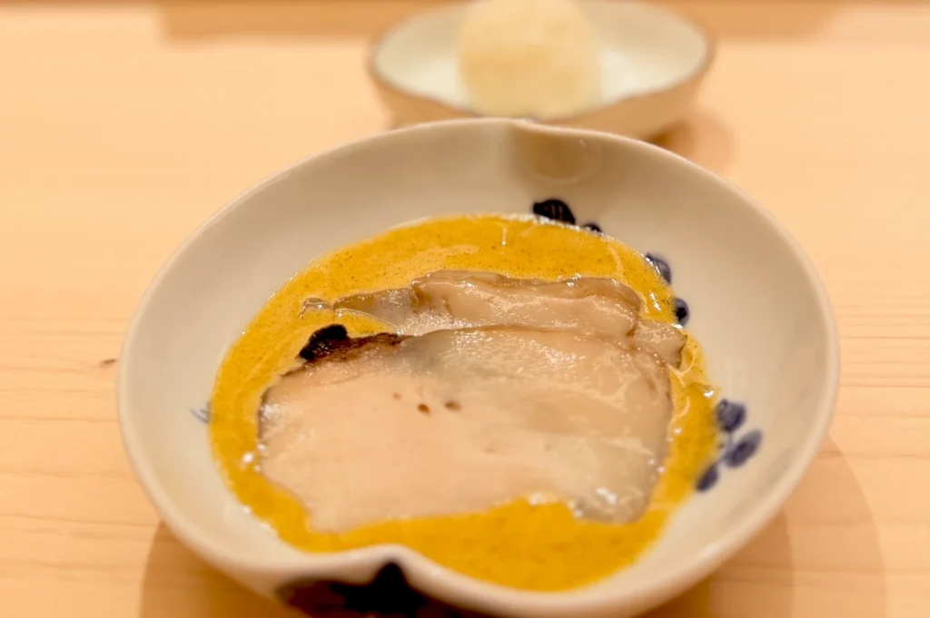 A delicate seafood dish featuring tender slices of abalone in a rich, savory yellow sauce of monkfish liver served in a patterned ceramic bowl at Sushi Sakuta.