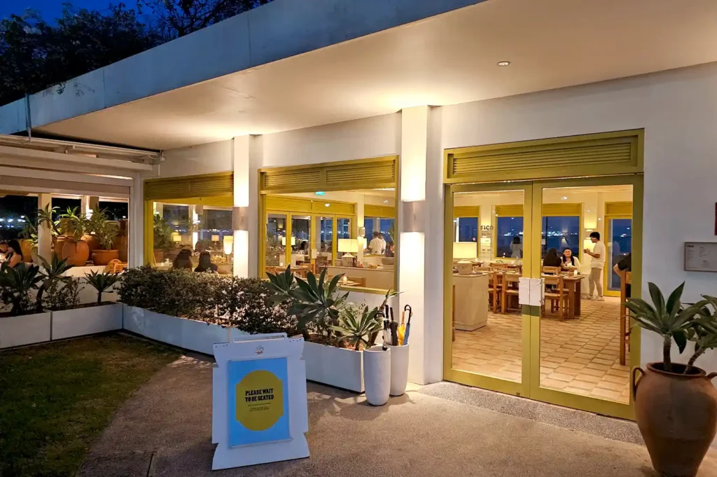 he warm, illuminated entrance and cozy indoor dining area of Fico Singapore at night, featuring glass doors, yellow window frames, and rustic potted plants.