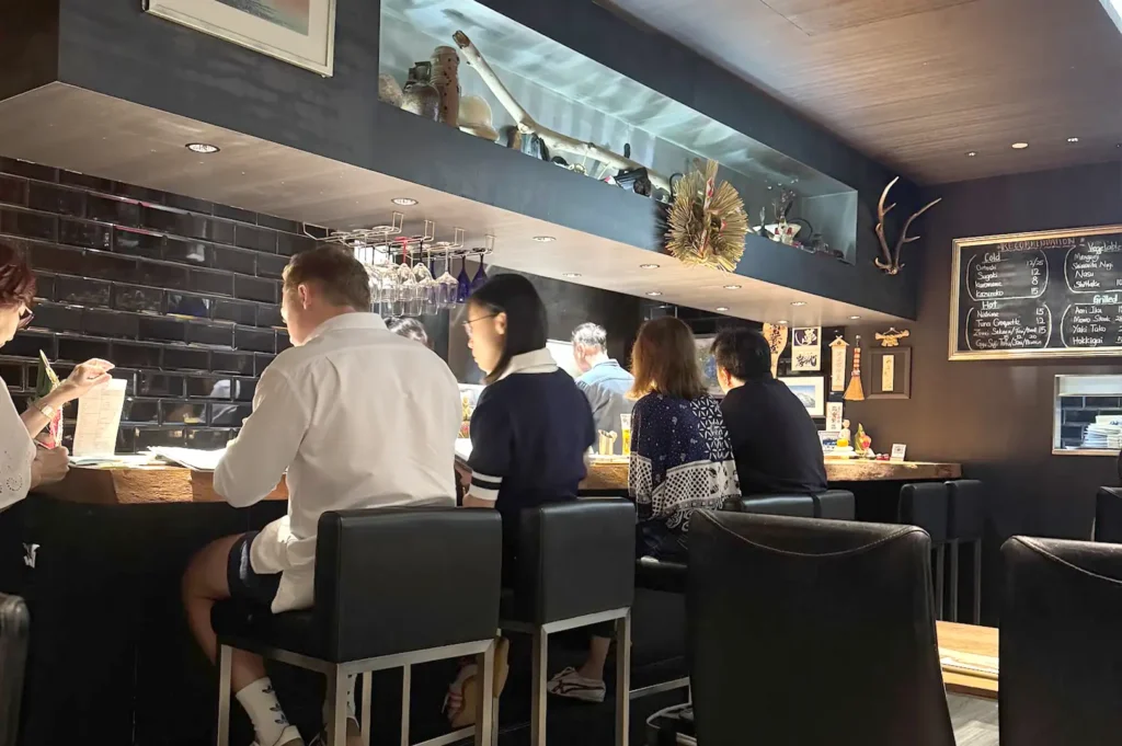 Diners seated along the sleek wooden counter at Kazu Sumiyaki Singapore, watching the live charcoal grilling action during a busy yakitori night.