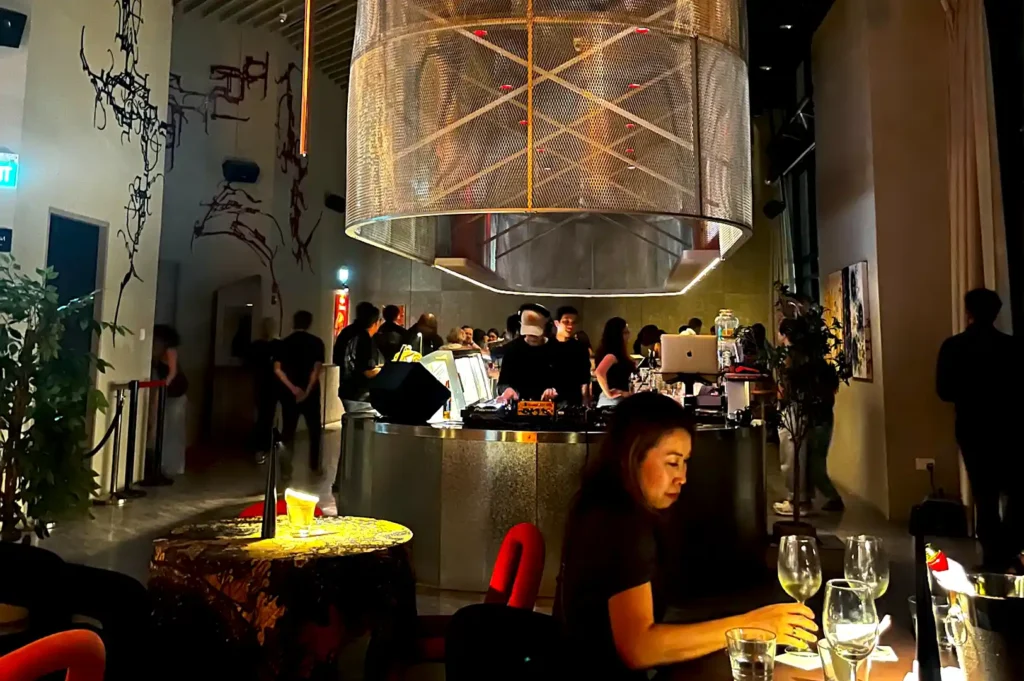 A vibrant evening scene inside Rasa Space at Republic Plaza, featuring patrons dining beneath a large metallic sculptural centerpiece and a live DJ station.