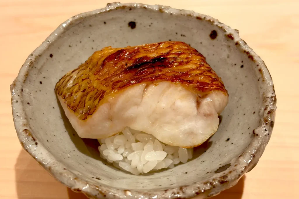 A piece of perfectly grilled, golden-brown fish served over a small bed of seasoned sushi rice in a textured ceramic bowl at Sushi Sakuta.