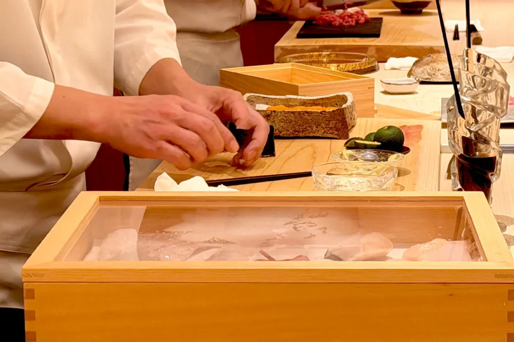 Close-up of a master chef's hands carefully shaping nigiri sushi behind a wooden counter equipped with fresh ingredients and traditional wooden neta boxes at Sushi Sakuta.