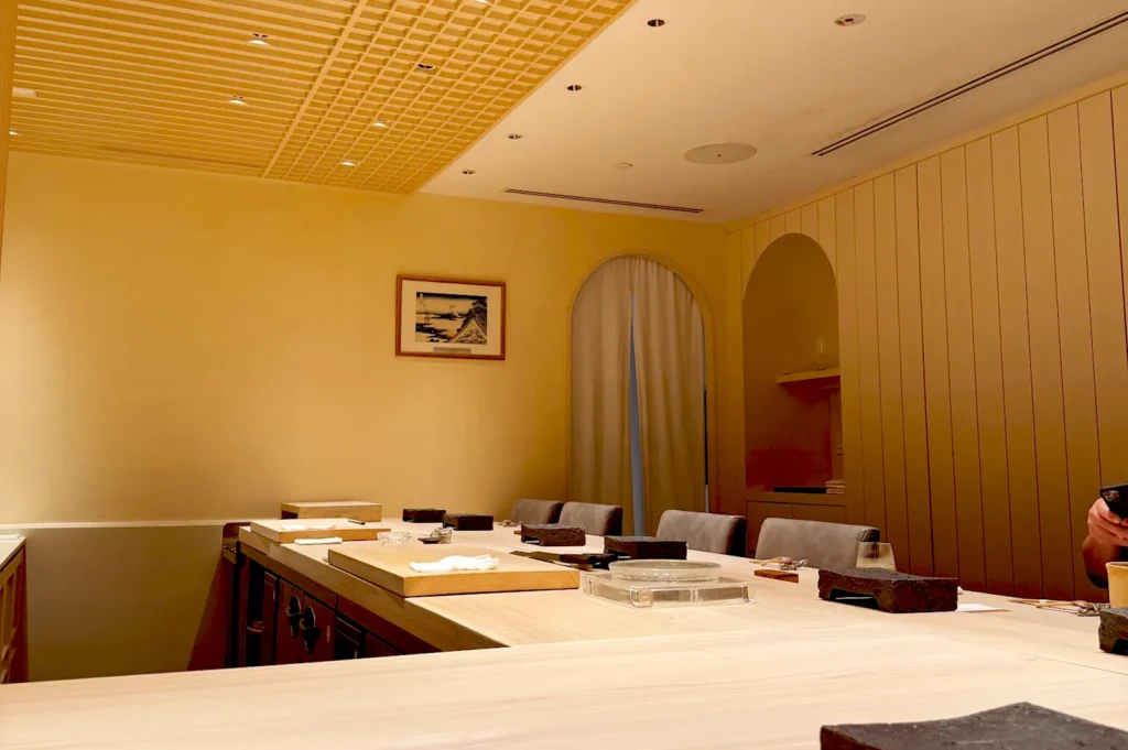 The minimalist and elegant dining interior of Sushi Sakuta, featuring light wood countertops, subtle lighting, warm-toned walls, and comfortable seating for an exclusive omakase experience.