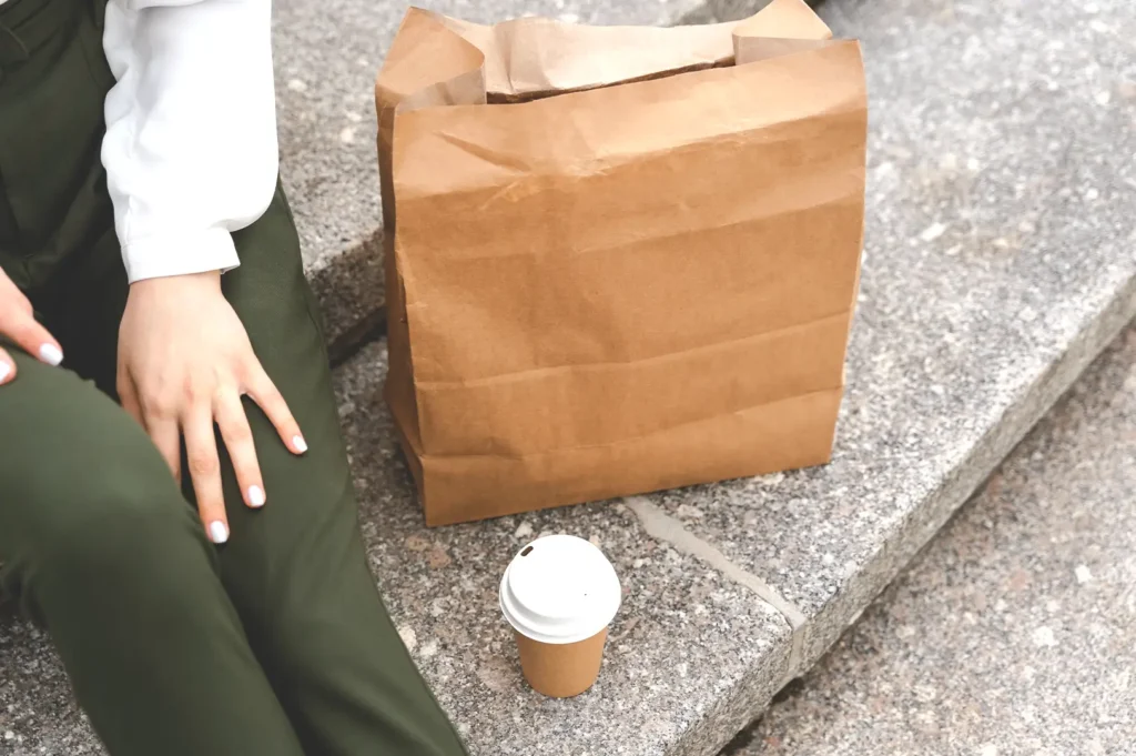 A person sitting on outdoor stone steps next to a large brown paper grocery bag and a takeaway coffee cup, reflecting the habit of carrying extra snacks, drinks, and emergency supplies.