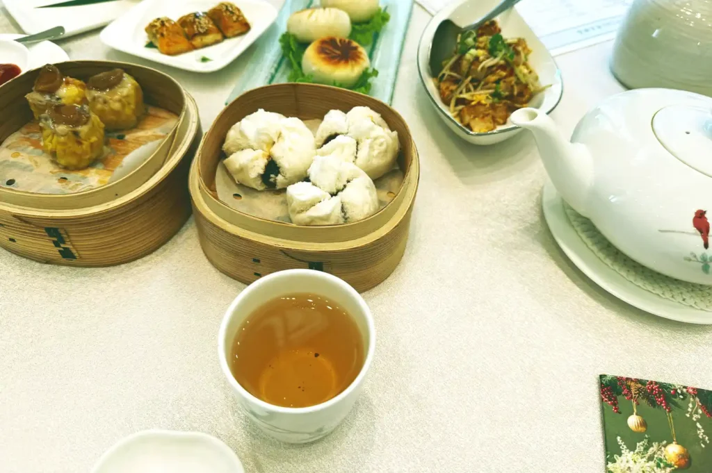 A full table spread of freshly steamed dim sum dishes, including bamboo baskets of fluffy char siu bao and siew mai, small plates of appetizers, a teapot, and a cup of Chinese tea, capturing the classic polite moment when everyone waits for someone else to start eating.