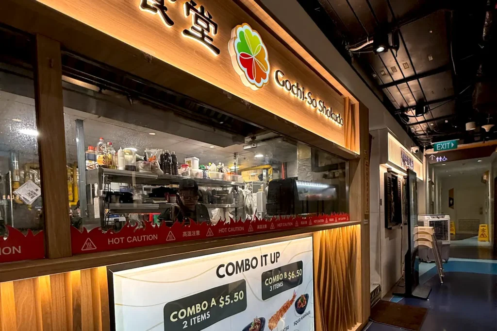 Storefront exterior of Gochi-So Shokudo at One Raffles Place featuring a wooden sign, warm lighting, and a view of the kitchen.