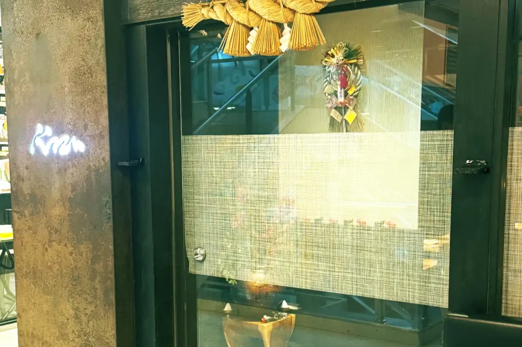 A sleek glass entrance of Kazu Sumiyaki at Cuppage Plaza, featuring traditional Japanese New Year ornaments (shimekazari) and a glowing neon sign.