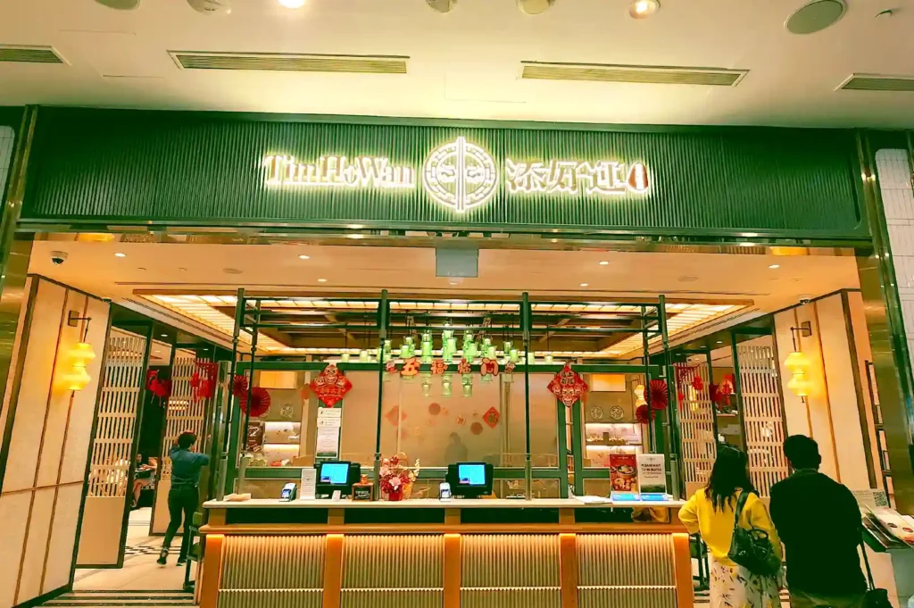 Storefront entrance of Tim Ho Wan PEAK at Marina Bay featuring a sleek green and wood design with reception counters.