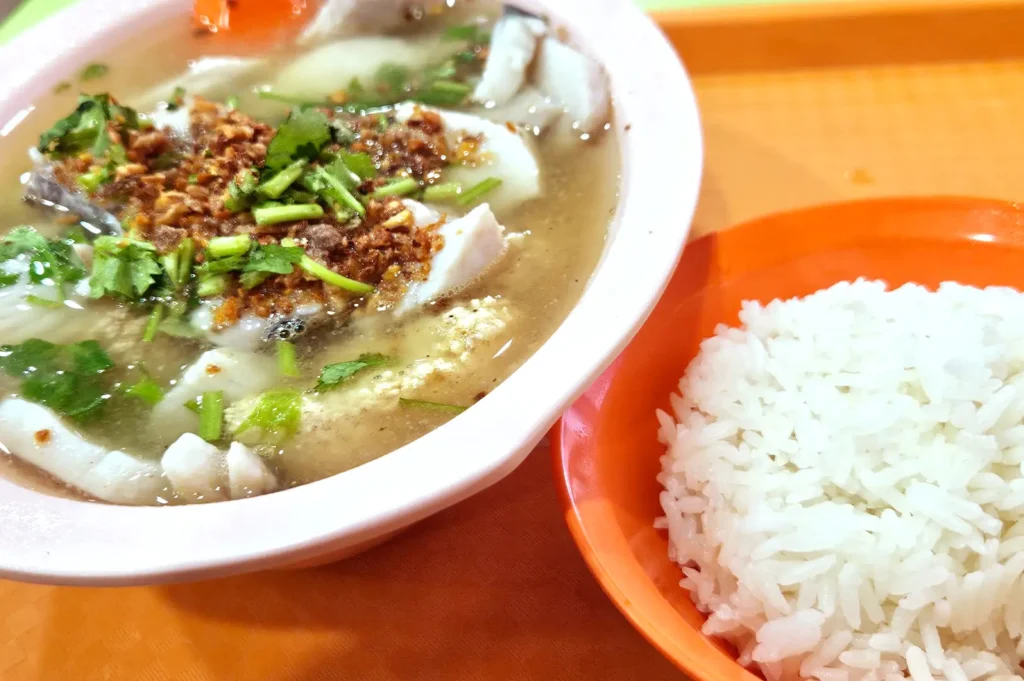 A bowl of clear soup topped with chopped green herbs and crispy fried garlic, served alongside a plate of steamed white rice on an orange table.
