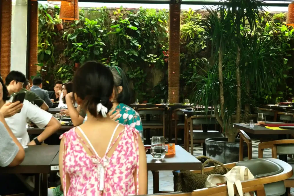 A lush, plant-filled restaurant interior with diners seated at tables, reflecting on the cultural expectations and culinary standards of older Singaporeans.