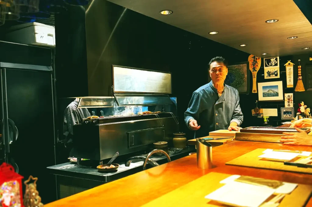 A chef attentively grilling skewers over a traditional charcoal sumiyaki station behind the wooden counter at Kazu Sumiyaki.