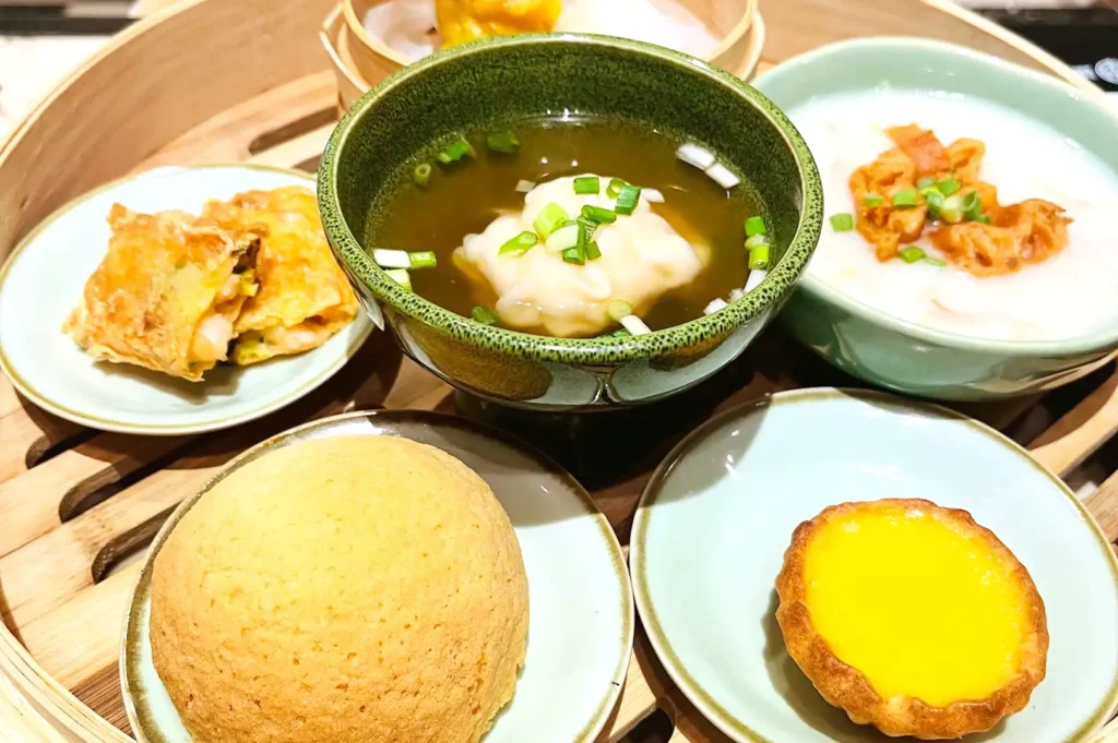 A wooden tray showcasing a spread of Tim Ho Wan signatures including baked BBQ pork buns, egg tarts, wonton soup, and congee.