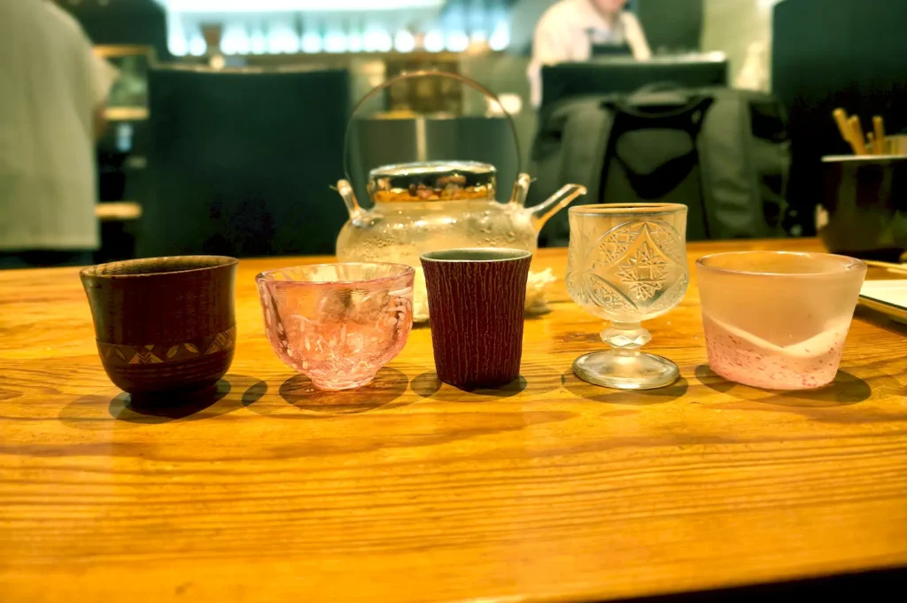 A neat alignment of various handcrafted Japanese ceramic cups, glassware, and a glass teapot resting on a wooden table.