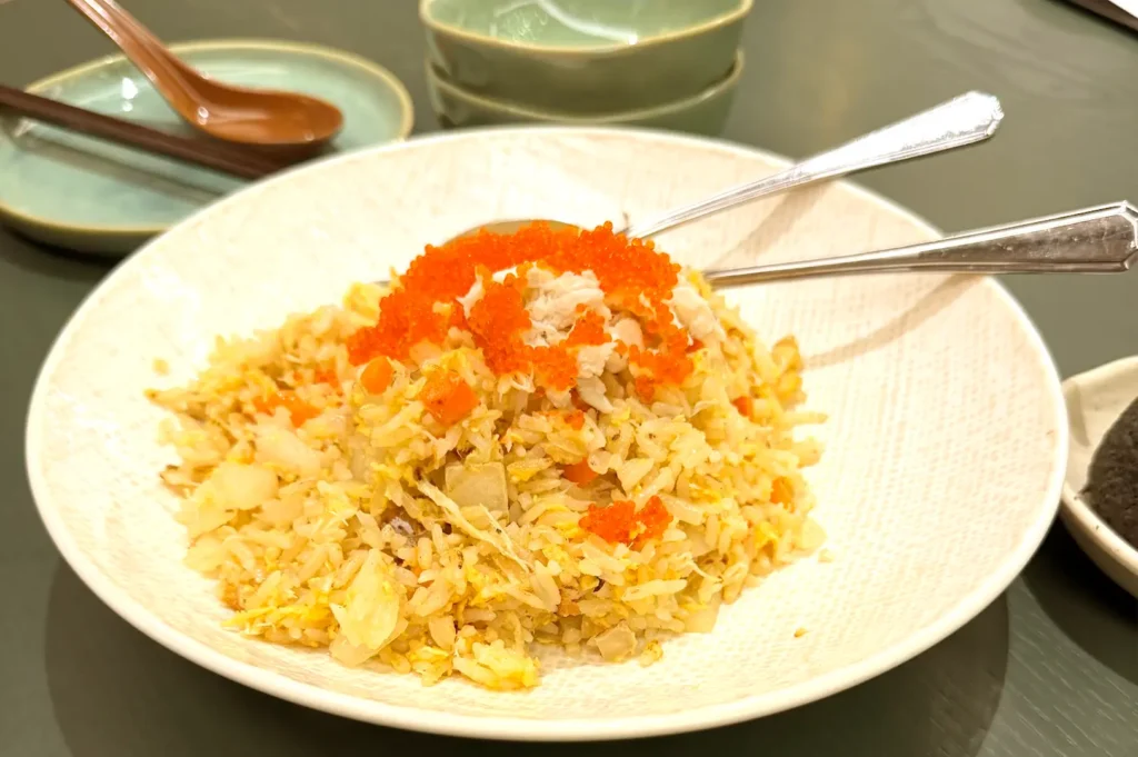 A white serving bowl of signature fried rice topped with bright orange fish roe, served with spoons and side dishes.