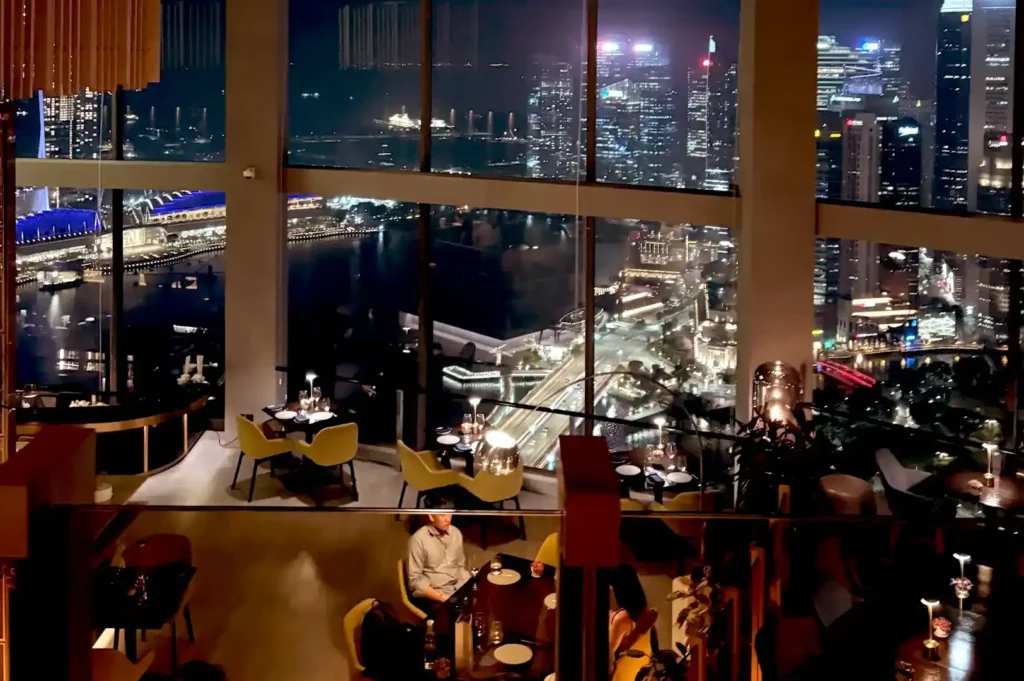 A stunning nighttime interior view of JAAN By Kirk Westaway, highlighting elegant dining tables overlooking floor-to-ceiling windows with a panoramic view of the glittering city skyline and waterfront.