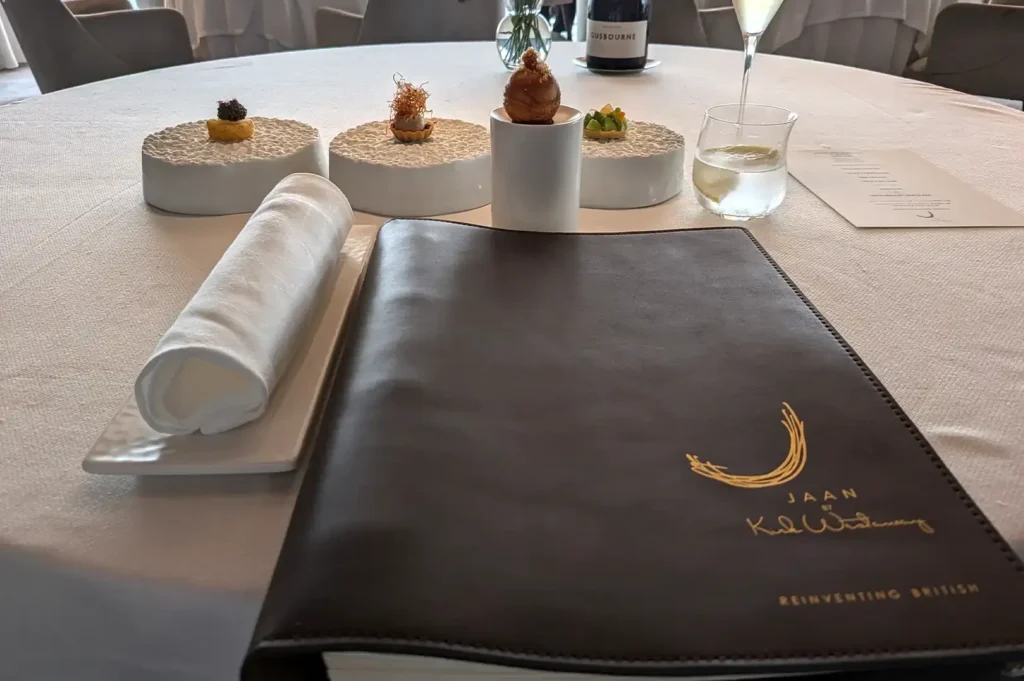A finely set dining table at JAAN By Kirk Westaway, showcasing a leather menu book with gold branding, a rolled white napkin, water glass, and an array of intricate canapés on textured white plates.