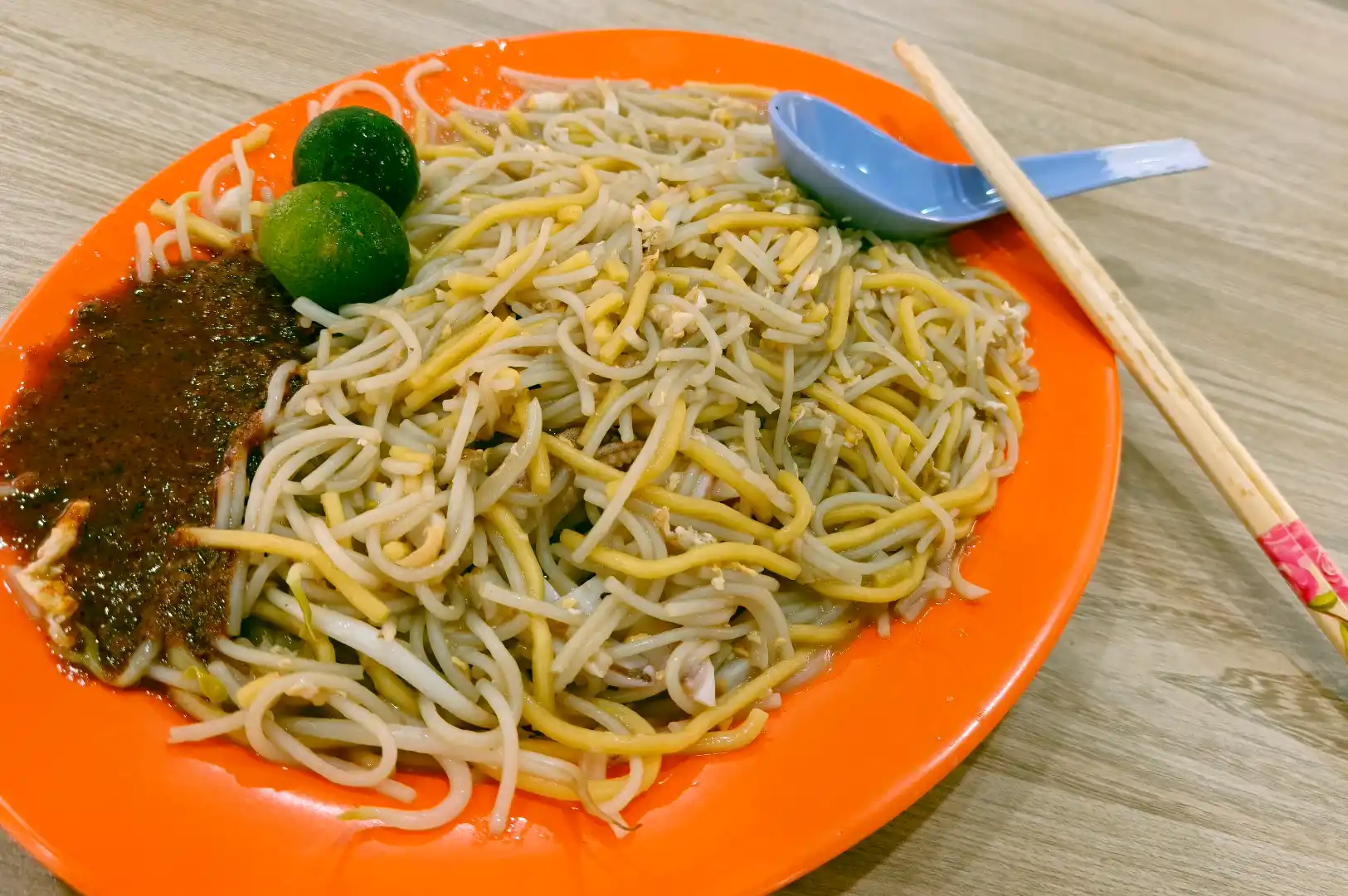 Overhead shot of a classic plate of Singapore Hokkien mee stir-fried with yellow noodles, thin bee hoon, calamansi limes, and a side of spicy sambal chili on an orange oval plate.