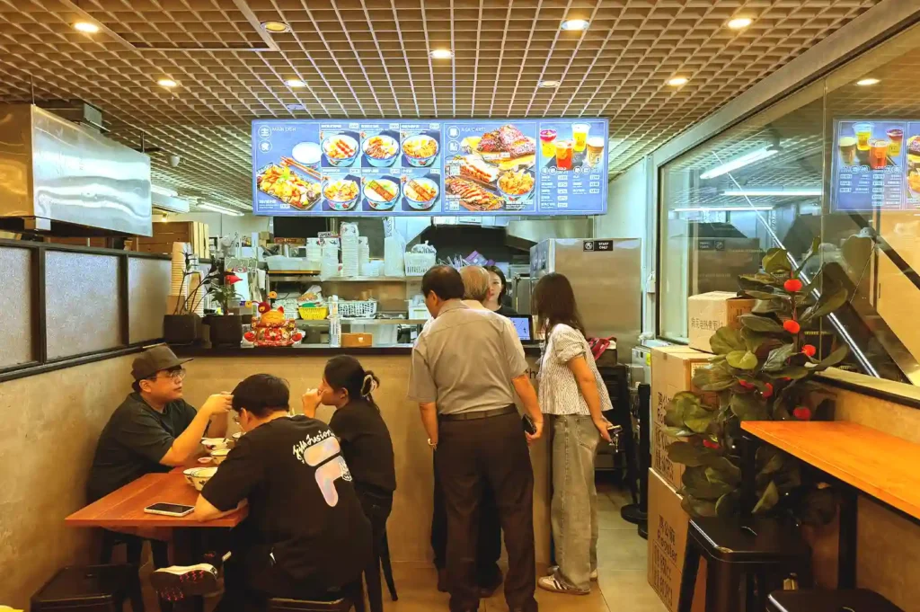The cozy interior of a casual Singaporean noodle eatery with patrons seated at wooden tables below an illuminated overhead menu board, capturing a hidden culinary gem saved on a personal bucket list.
