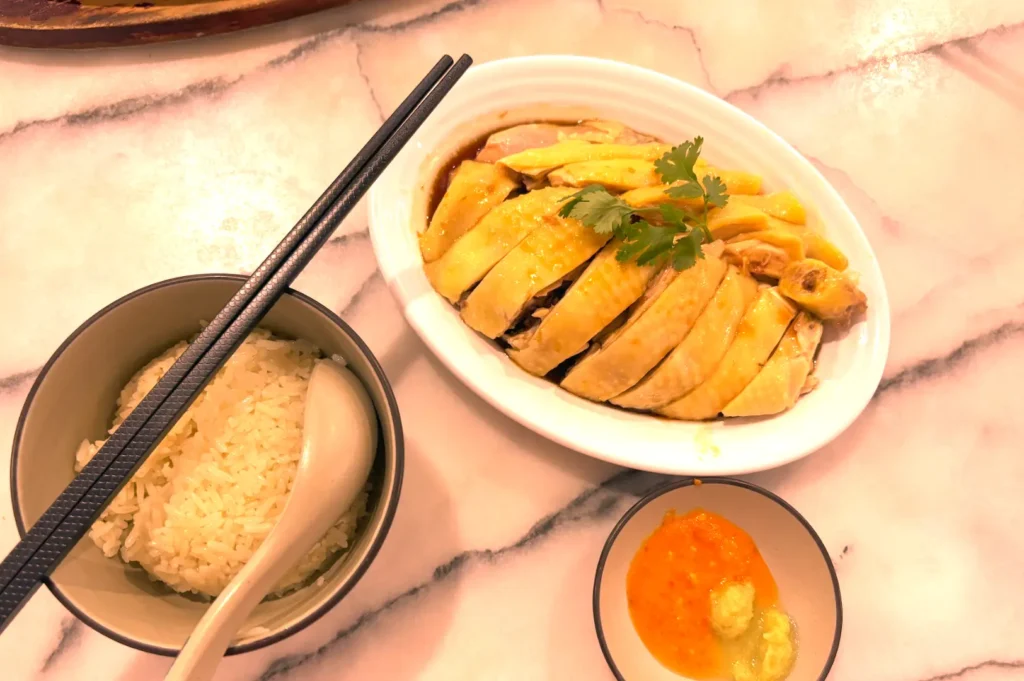 Overhead view of Hainanese poached chicken served on a white oval platter alongside a bowl of fragrant rice and chili ginger dipping sauce on a marble table, exemplifying meticulous culinary preparation and restaurant care.