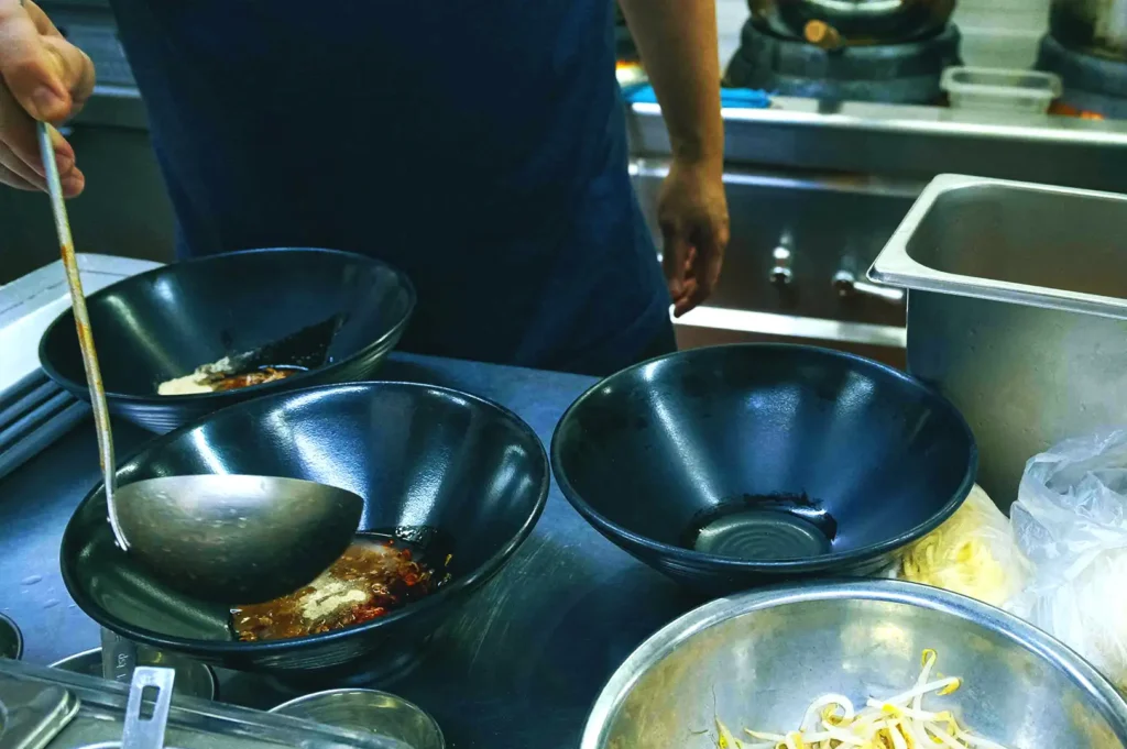 A chef preparing ingredients and broth bases in black bowls inside the bustling kitchen at Hup Lok.