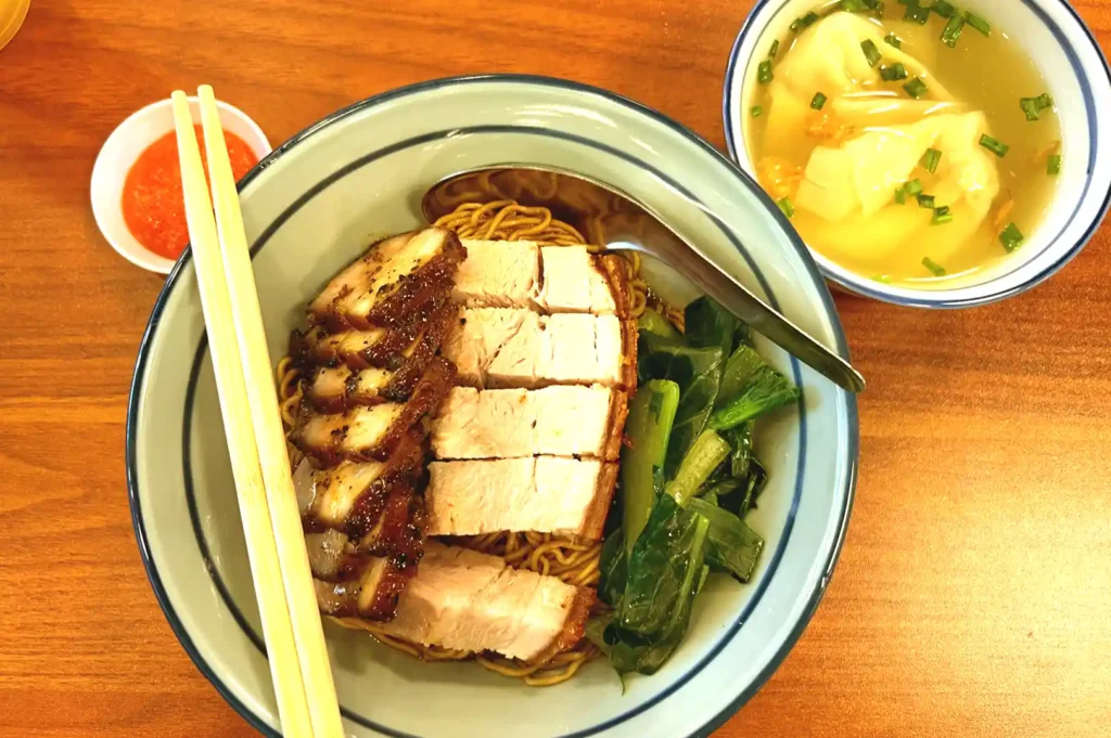A bowl of dry wanton noodles generously topped with caramelized char siu pork, crispy roasted pork belly, and leafy greens, served alongside a side of chili sauce and clear dumpling soup.