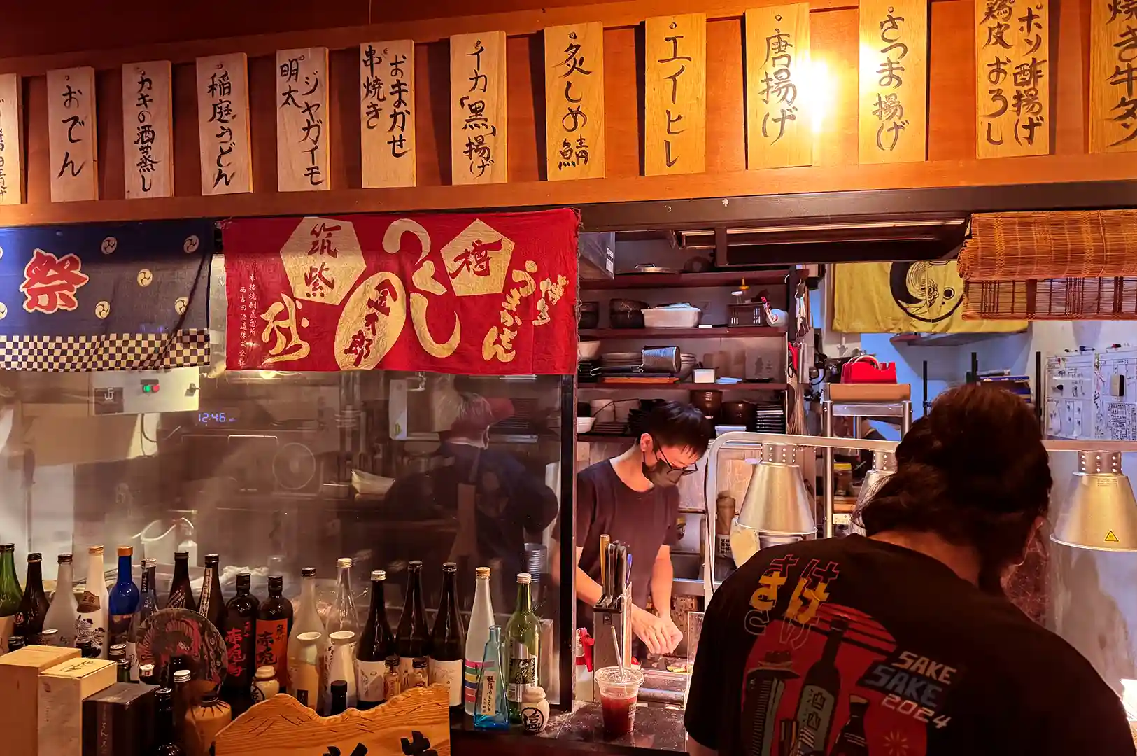 Chef preparing skewers in the open kitchen of ShuKuu Izakaya behind shelves lined with Japanese sake bottles and hanging menu boards.