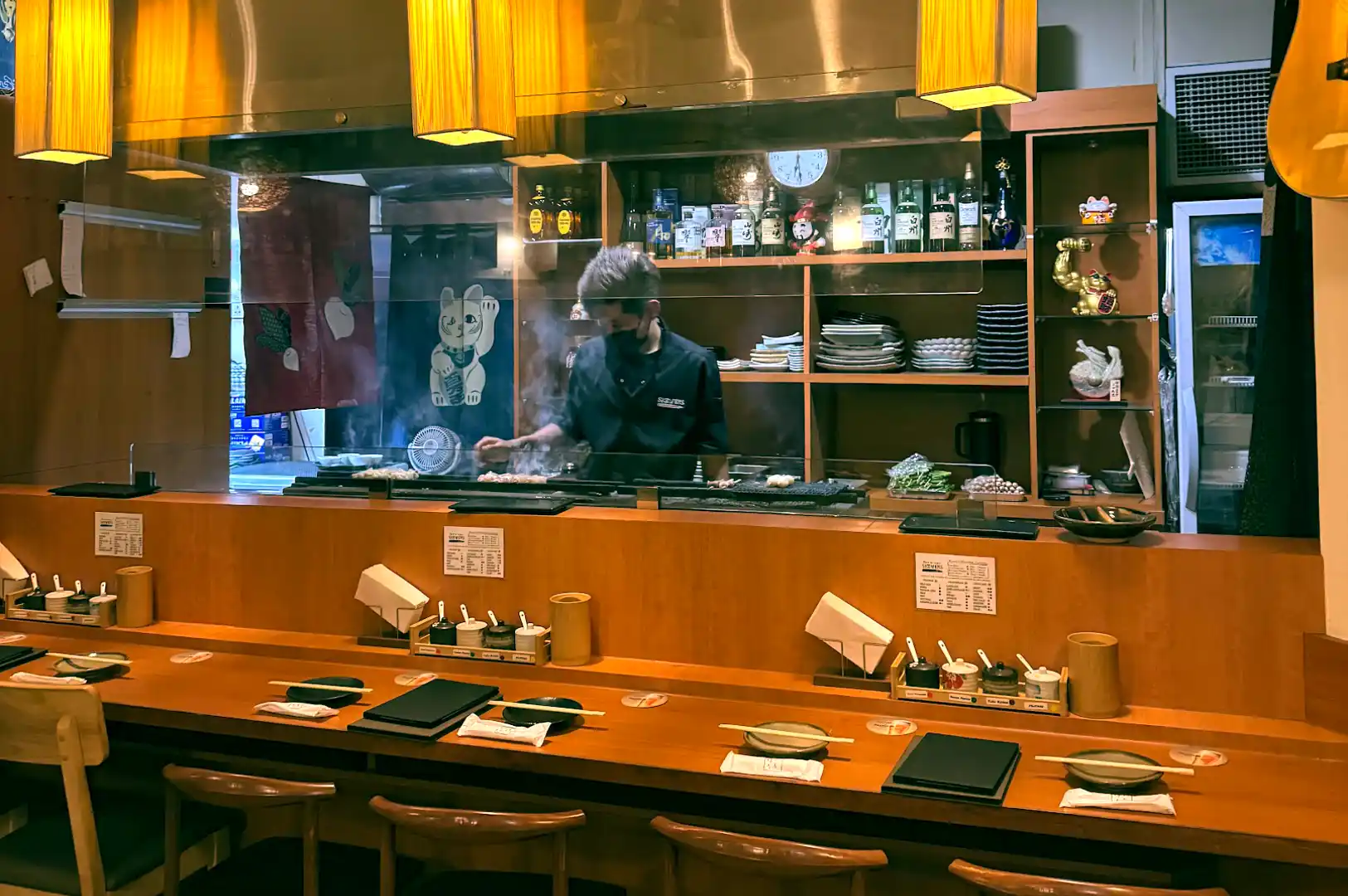 Chef grilling yakitori over charcoal behind a glass partition at the long wooden dining counter of Skewers Yakitori Izakaya.