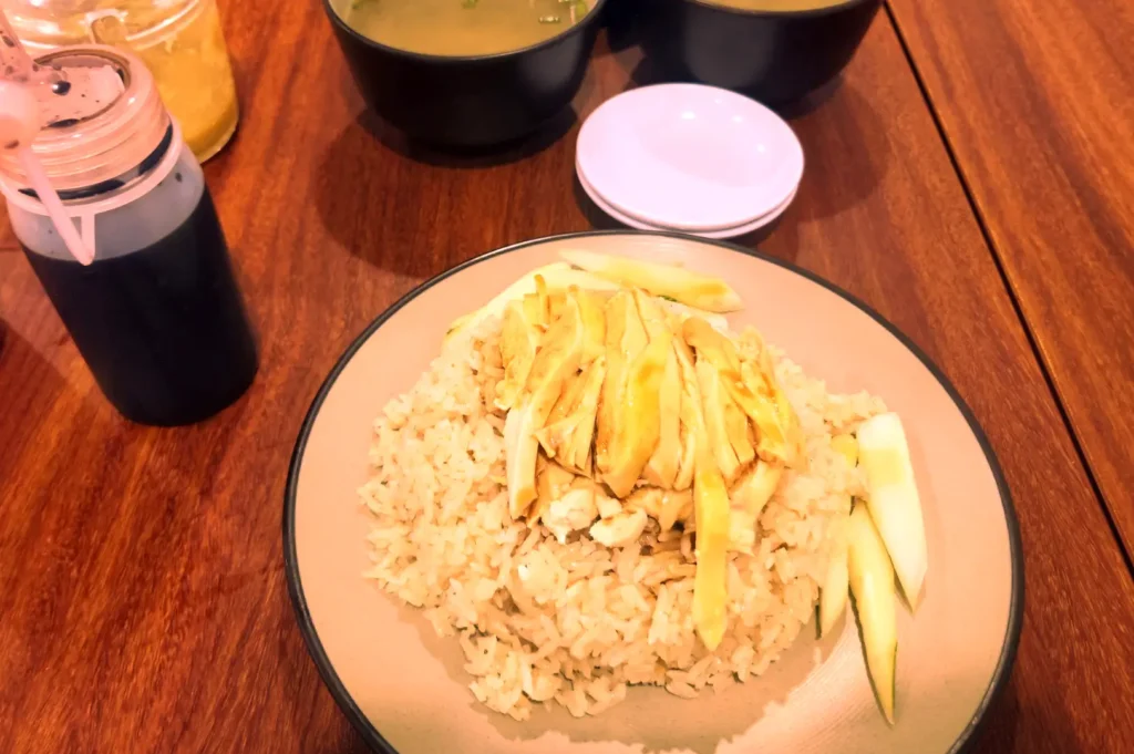 A plate of tender sliced poached chicken over fragrant rice garnished with cucumber slices, served next to dark soy sauce squeeze bottles and soup bowls on a wooden dining table.