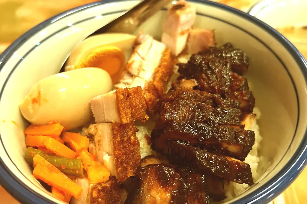A close-up of a rice bowl loaded with thick-cut roasted pork belly with crackling skin, dark glazed braised meat, pickled vegetables, and a soft-boiled ramen egg.