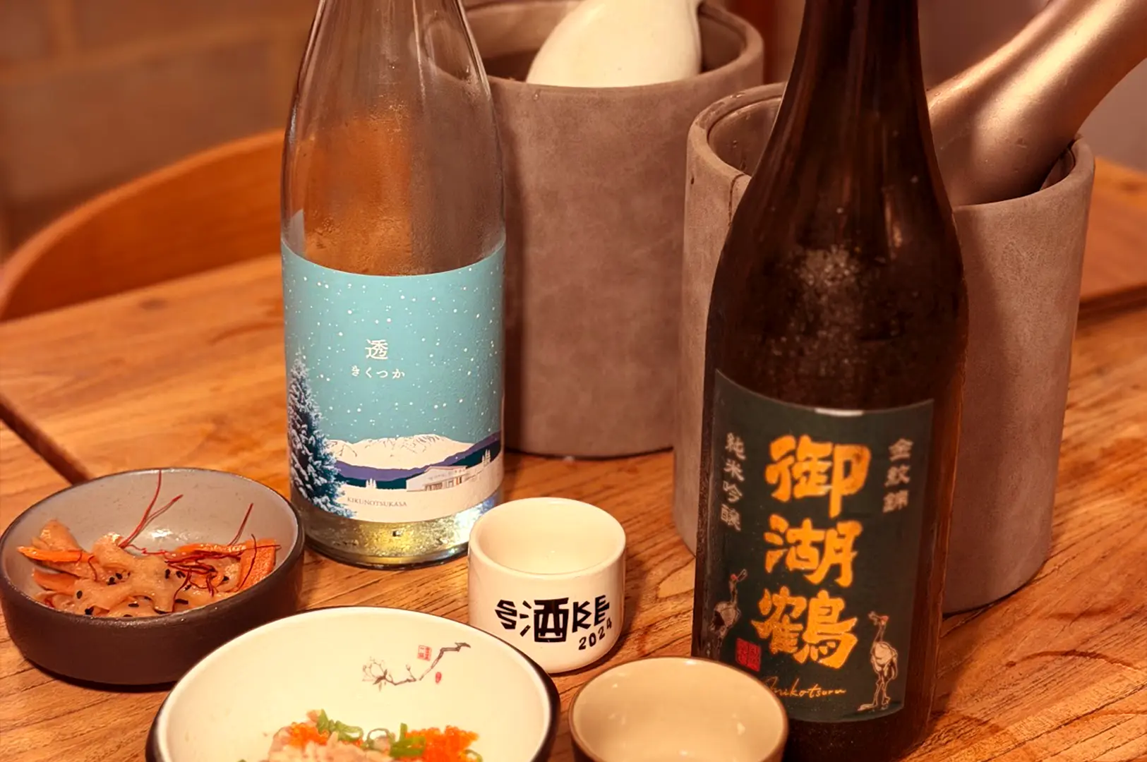 Chilled bottles of Japanese sake served on a wooden table alongside Ochoko sake cups and side dishes at ShuKuu Izakaya.