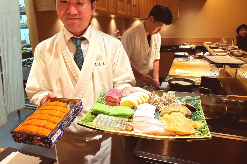 A smiling chef at Tenshima proudly showcasing a fresh platter of premium seasonal ingredients, including sea urchin (uni), abalone, wagyu beef, and seafood ready for tempura.