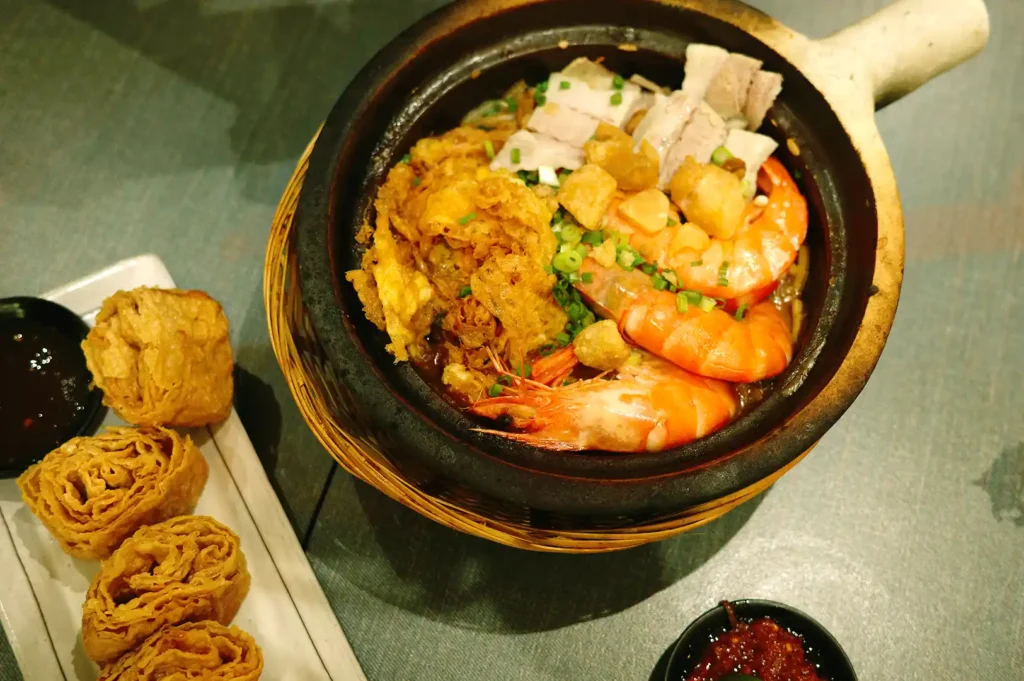 A hearty claypot dish from Hup Lok featuring plump prawns, sliced pork belly, crispy egg floss, and rich noodles, served alongside crispy bean curd rolls.