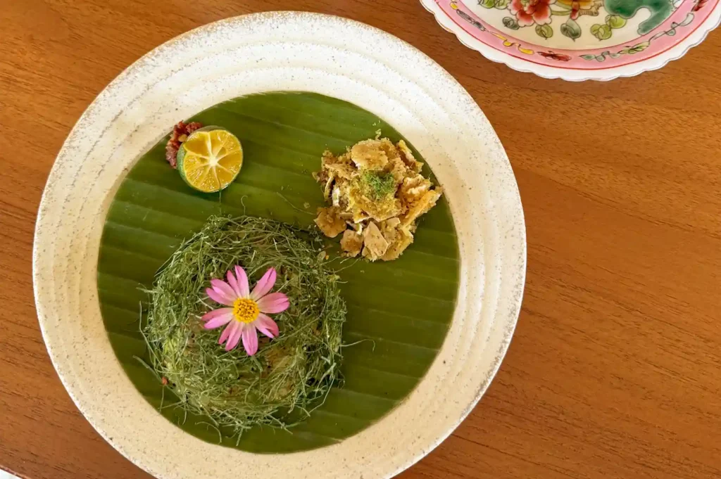 A beautifully plated dish highlighting Buak Keluak inspiration as an ingredient, served on a banana leaf with delicate herbs and edible flowers.