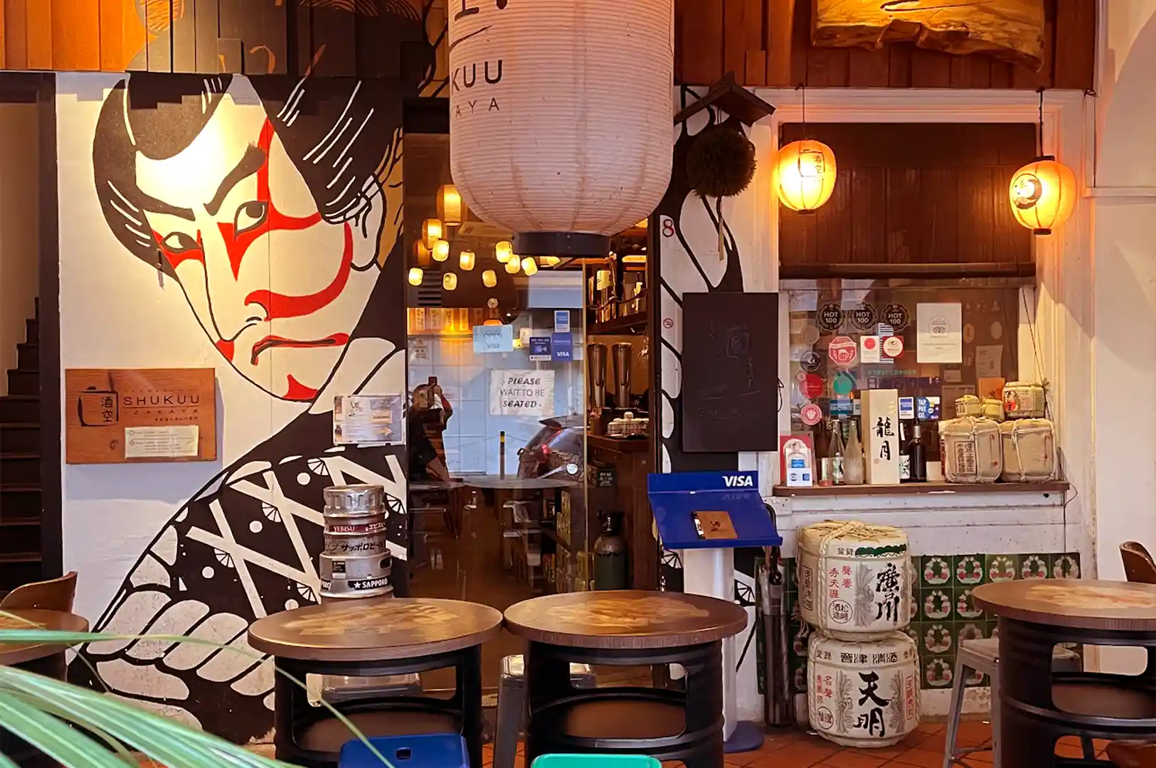 Exterior entrance of ShuKuu Izakaya showing outdoor seating tables, traditional sake barrels, paper lanterns, and Kabuki artwork.