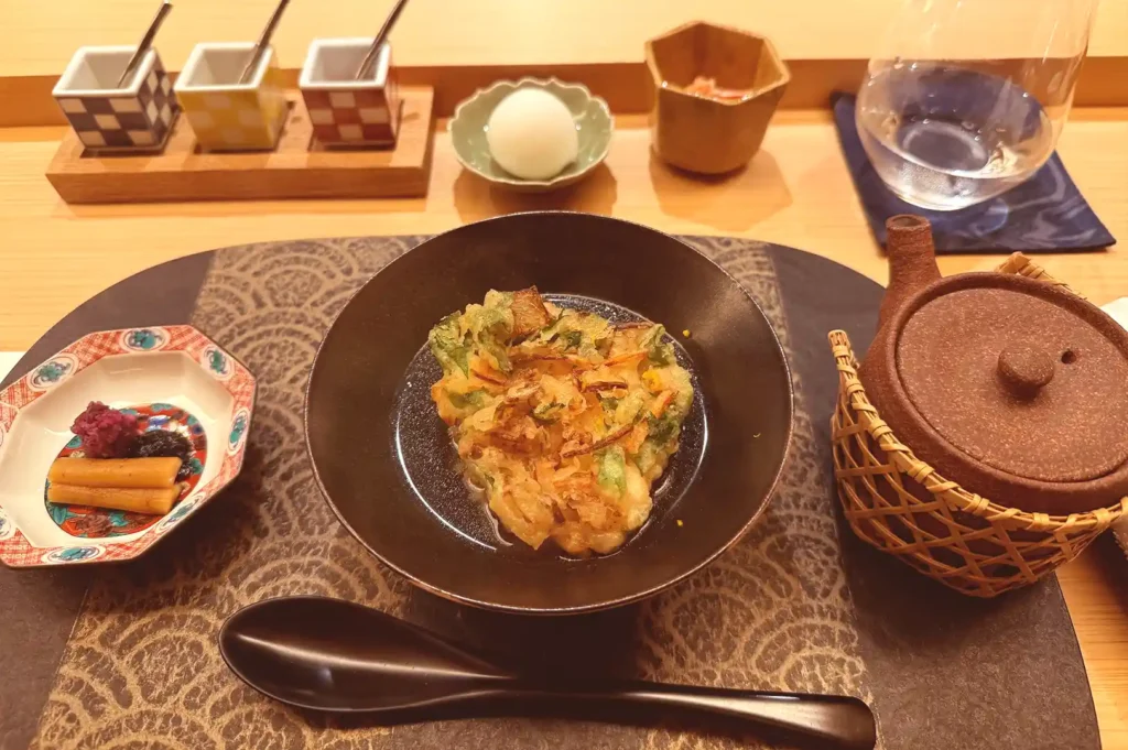 An exquisite course from the Tenshima tempura omakase menu featuring delicately battered and fried seasonal items served in a traditional black bowl with condiments and a teapot.