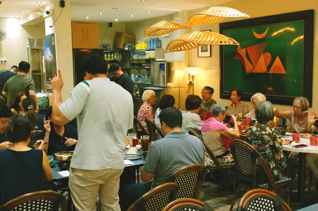 A lively and warm dining room scene at Hup Lok featuring groups of families and friends enjoying comforting seafood dishes beneath modern leaf-shaped pendant lamps.