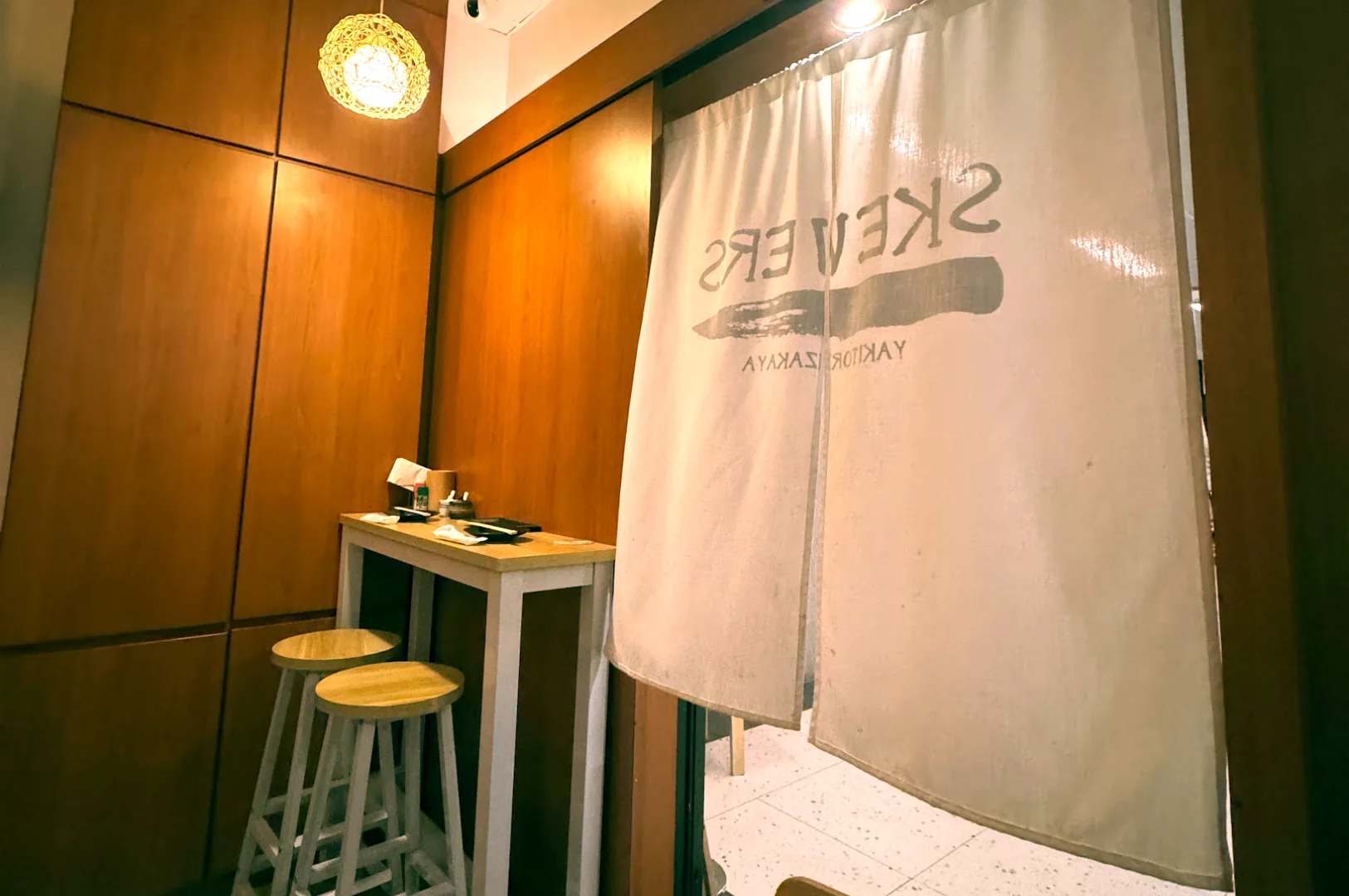 View looking past the white printed noren curtain into an intimate dining nook with wooden bar stools at Cuppage Plaza.
