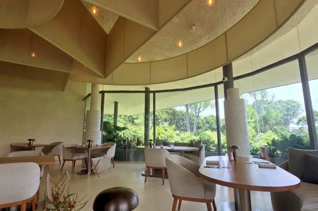 A panoramic interior view of Pangium's sophisticated dining room framed by curved glass windows that merge the garden landscape with the restaurant's heritage-inspired aesthetic.