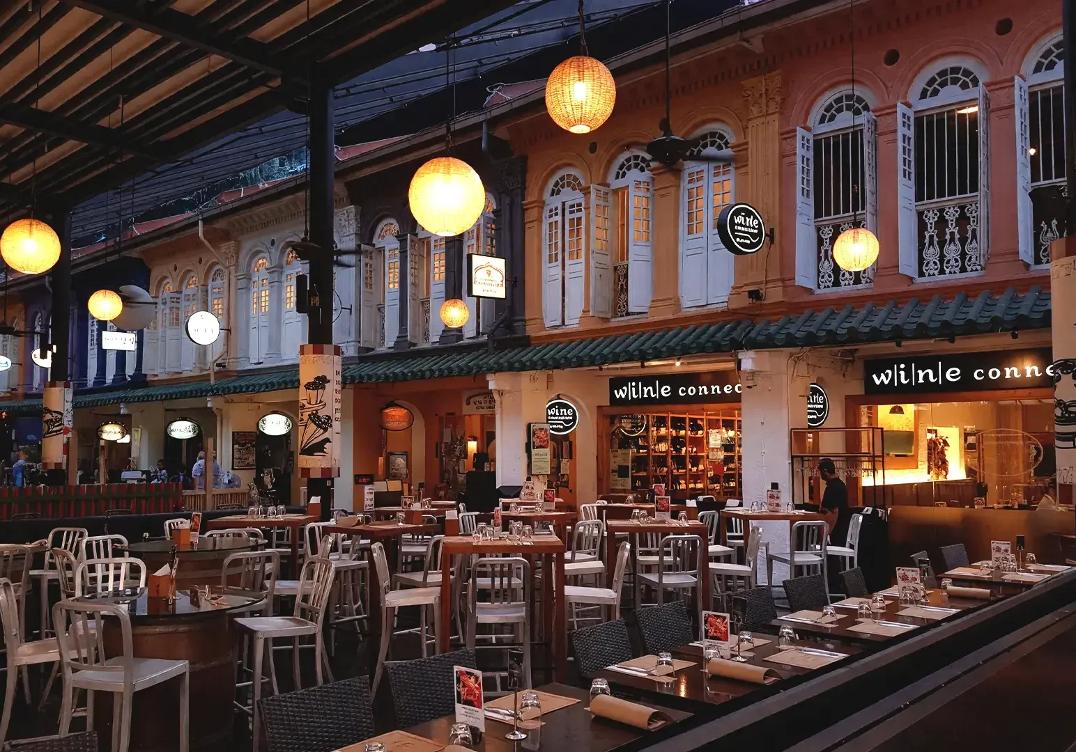 A vibrant evening view of an outdoor dining area at Somerset for late-night feasts, featuring rows of dining tables set with plates and silverware, surrounded by colorful, illuminated shophouse facades. The space is warmly lit by hanging lanterns and ambient lights, inviting friends for food. 