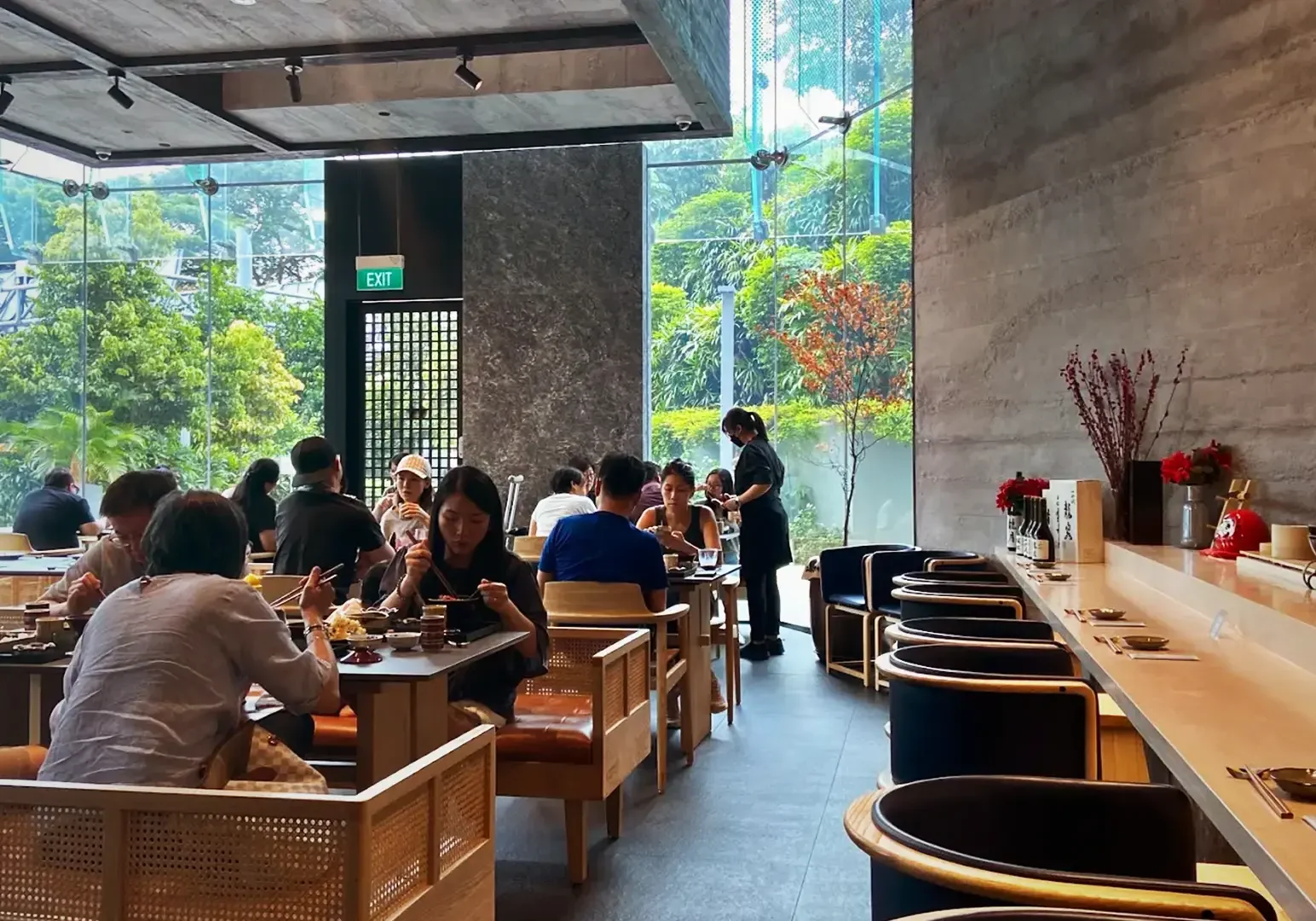 Patrons enjoying a premium lunch omakase experience inside the modern, glass-walled dining area at Ganko Sushi, featuring minimalist wooden furniture and lush garden views.